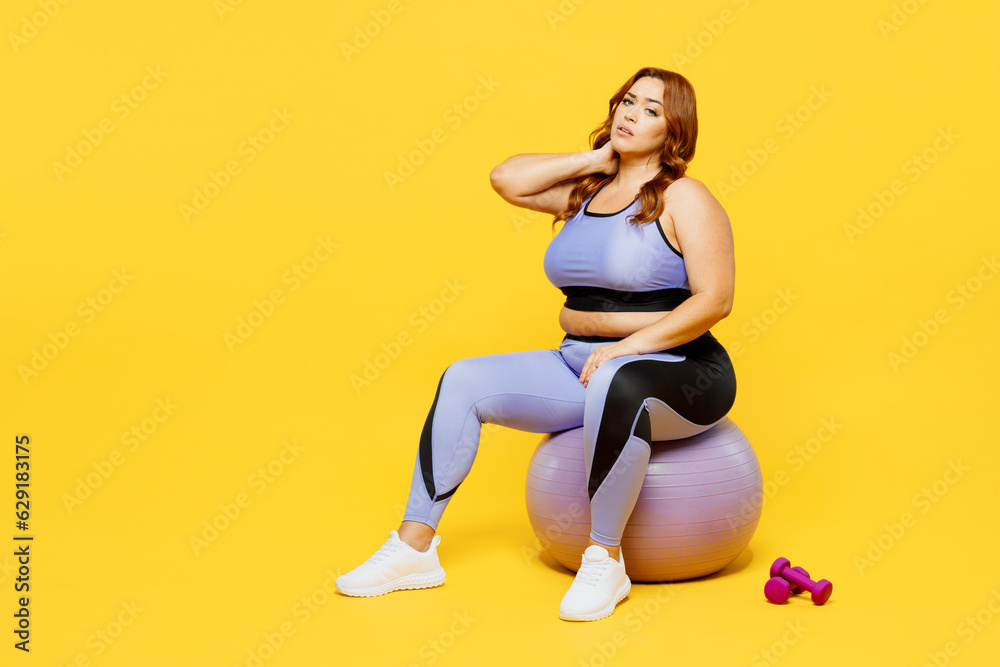 Full body young chubby plus size big fat fit woman wear blue top warm up training sit on fit ball hold neck suffer from pain isolated on plain yellow background studio home gym. Workout sport concept. Full body young chubby plus size big fat fit woman wear blue top warm up training sit on fit ball hold neck suffer from pain isolated on plain yellow background studio home gym. Workout sport concept.