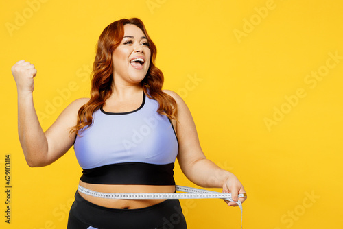 Young overweight plus size big fat fit woman wearing blue top warm up training hold measure tape on waist do winner gesture isolated on plain yellow background studio home gym. Workout sport concept.