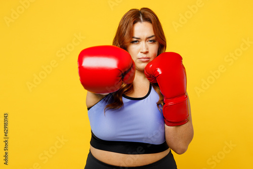Young confident strict chubby overweight plus size big fat fit woman in blue top red gloves warm up train boxing look camera isolated on plain yellow background studio home gym Workout sport concept © ViDi Studio