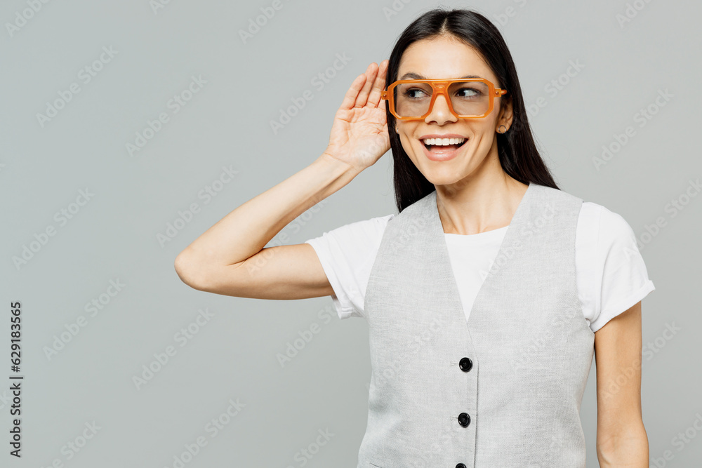 Young smiling fun curious nosy latin woman she wears white t-shirt gray vest glasses try to hear you overhear listening intently isolated on plain grey background studio portrait. Lifestyle concept. Young smiling fun curious nosy latin woman she wears white t-shirt gray vest glasses try to hear you overhear listening intently isolated on plain grey background studio portrait. Lifestyle concept.