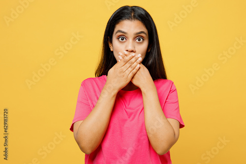 Young scared fearful frightened shocked sad Indian woman wearing pink t-shirt casual clothes look camera cover mouth with hand isolated on plain yellow background studio portrait. Lifestyle concept.
