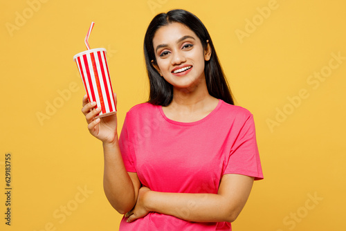 Young cool happy Indian woman wearing pink t-shirt casual clothes hold in hand cup of soda pop cola fizzy water looking camera isolated on plain yellow background studio portrait. Lifestyle concept.