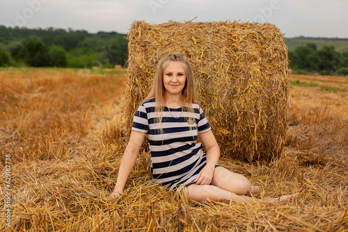 a long-haired blonde girl in a light summer dress is leaning against a haystack in a wheat field 