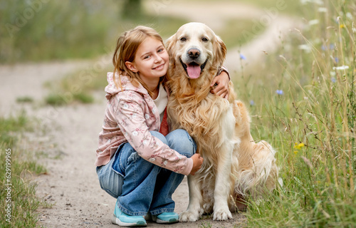 Preteen girl with golden retriever dog sitting at nature and looking back. Cute child kid hugging purebred pet doggy in park at summer