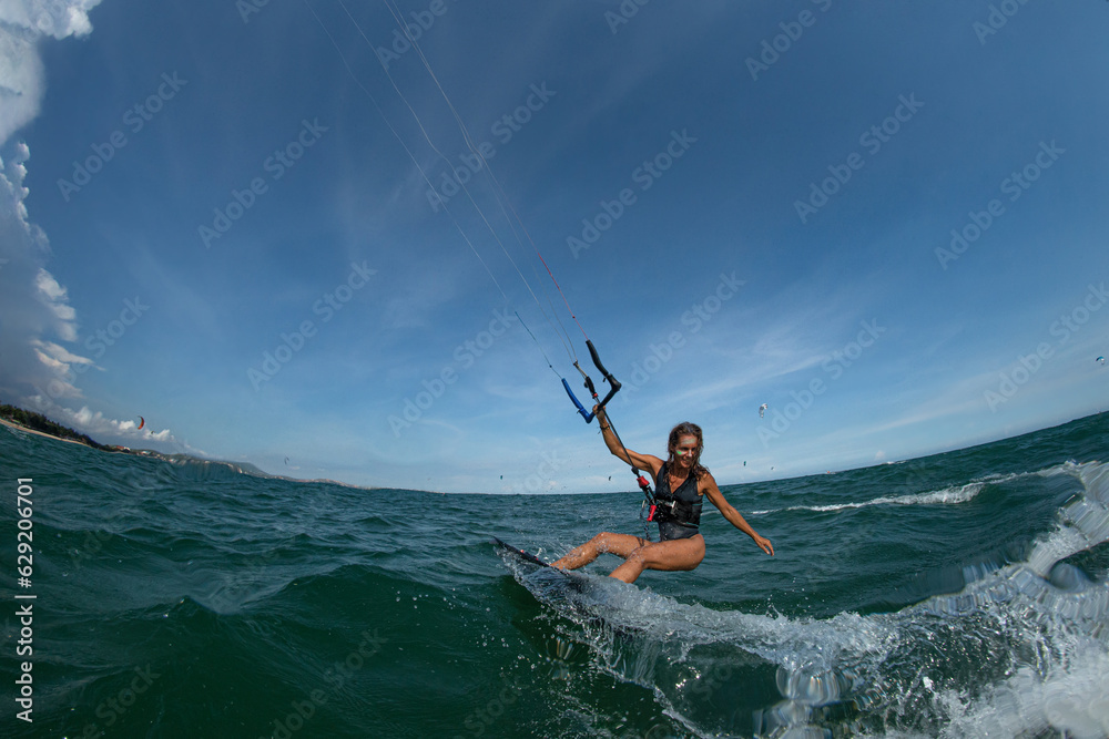 Kitesurfing girl in black sexy swimsuit with kite in sky on board in blue sea riding waves with water splash. Recreational activity, water sports, action, hobby and fun in summer time. Kitesurfing girl in black sexy swimsuit with kite in sky on board in blue sea riding waves with water splash. Recreational activity, water sports, action, hobby and fun in summer time.