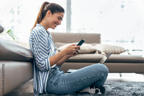 Foto Shot of pretty young woman using her mobile phone while working with laptop sitting on the floor at home