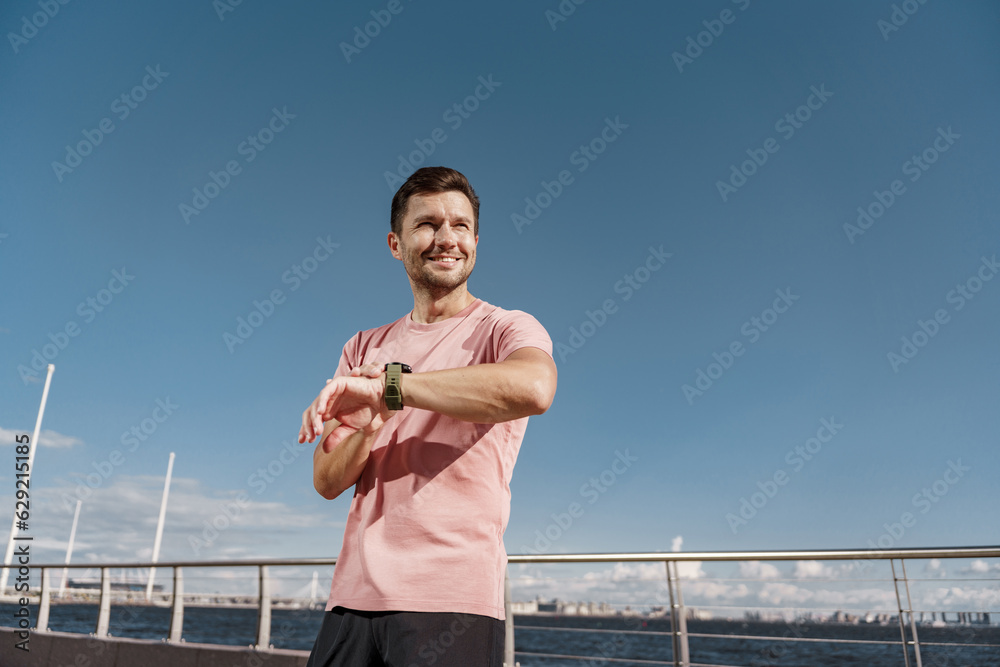 Using a smartwatch, a runner guy in fitness clothes looks at the time. A man does sports. A happy athlete is a person.