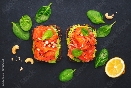 Avocado and salmon sandwich or toast on rye bread with spinach, crushed cashew nuts and sesame seeds, black table background, top view