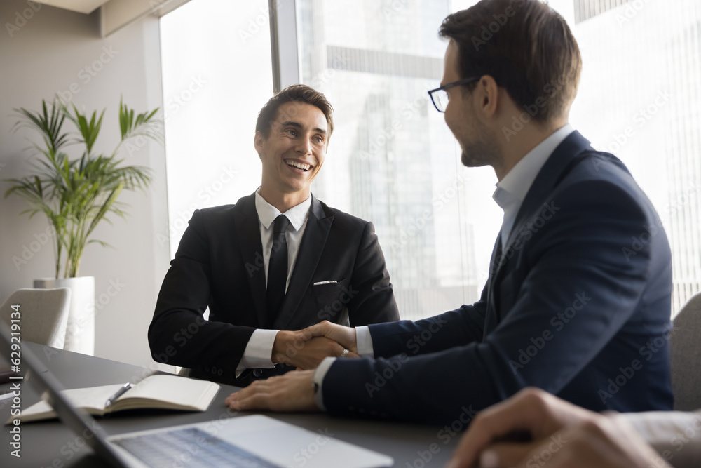 Two happy confident businessmen shaking hands, sitting at conference table, closing meeting, contract, successful project, talking, smiling, laughing, giving handshakes after job interview Two happy confident businessmen shaking hands, sitting at conference table, closing meeting, contract, successful project, talking, smiling, laughing, giving handshakes after job interview