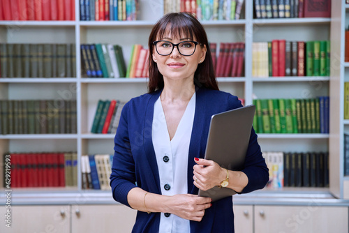 Portrait of confident female teacher holding laptop inside library
