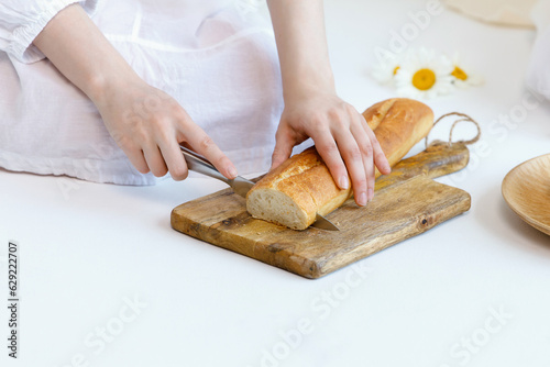 Girl cuts a loaf of bread with a knife on a wooden baking sheet on floor. Part of body