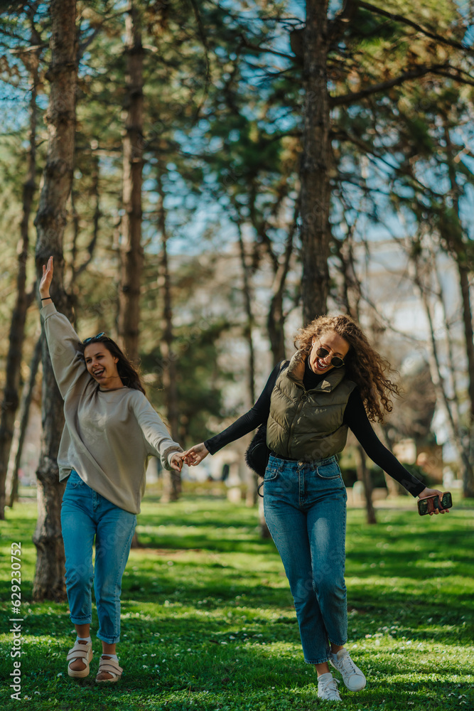Fototapeta premium Front view shot of two lovely, smiley girls hanging out in the park with spread arms while holding their hands