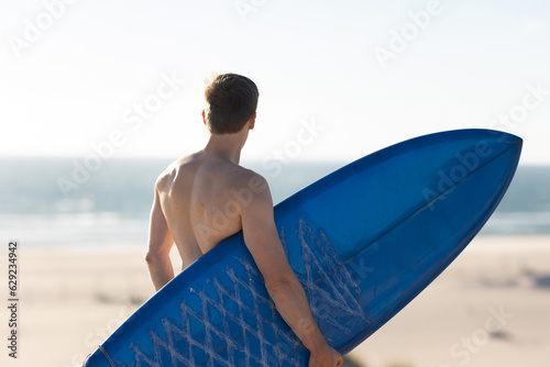A man with naked torso standing on the beach holding a blue surfboard - view from the back