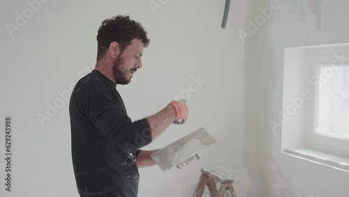 Caucasian working man preparing plaster to improve and remodel white surface on indoor drywalls in house construction site, handyman at work with palette knife, renovation project in domestic interior