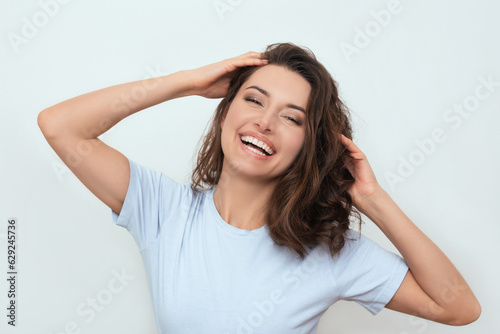 a photo of a positive young happy dark-haired Caucasian woman in a blue T-shirt, who smiling and touching her curly hair with her hands. portrait of a beautiful model. advertisement