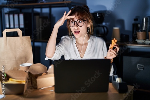 Young beautiful woman working using computer laptop and eating delivery food very happy and smiling looking far away with hand over head. searching concept.