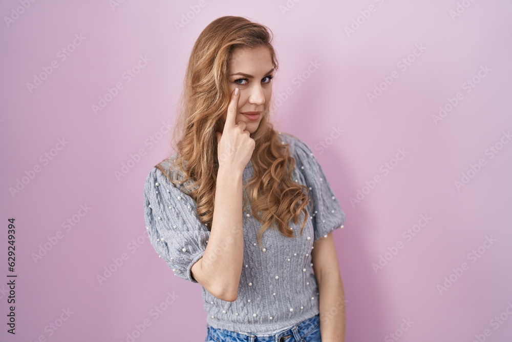 Beautiful blonde woman standing over pink background pointing to the eye watching you gesture, suspicious expression Beautiful blonde woman standing over pink background pointing to the eye watching you gesture, suspicious expression