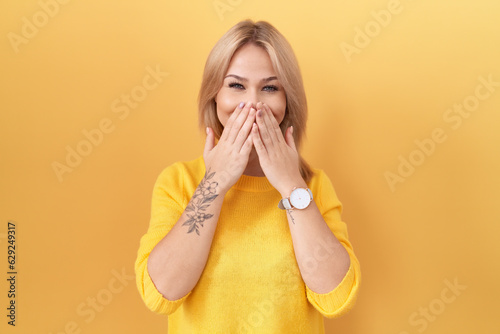 Young caucasian woman wearing yellow sweater laughing and embarrassed giggle covering mouth with hands, gossip and scandal concept photo