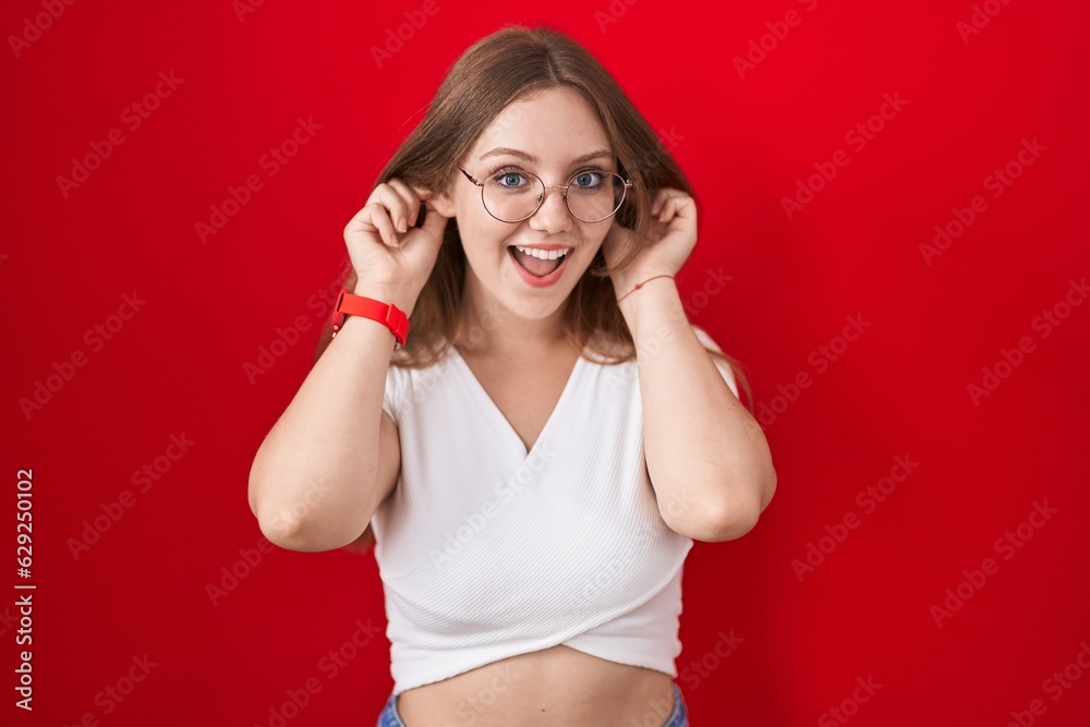 Young caucasian woman standing over red background smiling pulling ears with fingers, funny gesture. audition problem