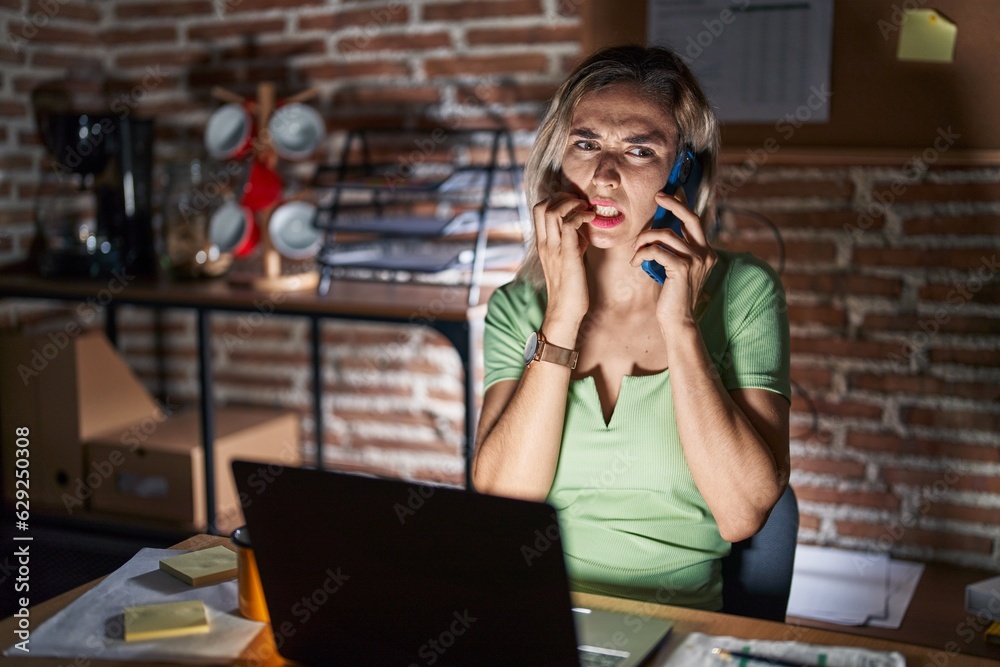 Young beautiful woman working at the office at night speaking on the phone looking stressed and nervous with hands on mouth biting nails. anxiety problem. Young beautiful woman working at the office at night speaking on the phone looking stressed and nervous with hands on mouth biting nails. anxiety problem.