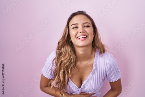 Young hispanic woman standing over pink background smiling and laughing hard out loud because funny crazy joke with hands on body.
