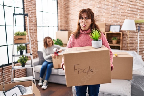Mother and daughter moving to a new home holding cardboard box sticking tongue out happy with funny expression. © Krakenimages.com