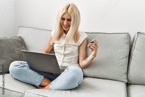 Young blonde woman shopping with laptop and credit card sitting on sofa at home