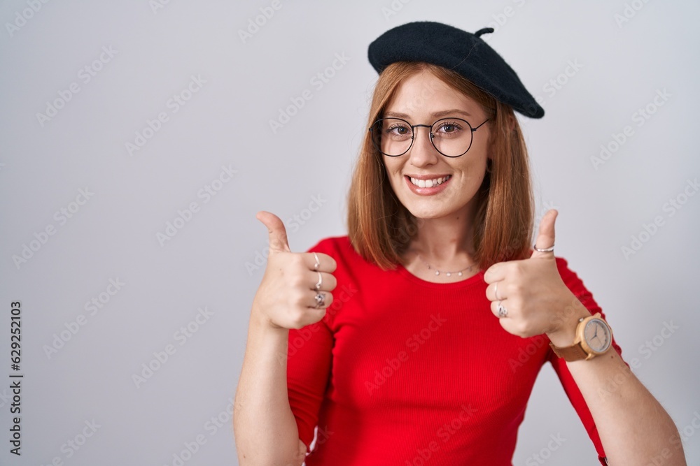 Young redhead woman standing wearing glasses and beret success sign doing positive gesture with hand, thumbs up smiling and happy. cheerful expression and winner gesture. Young redhead woman standing wearing glasses and beret success sign doing positive gesture with hand, thumbs up smiling and happy. cheerful expression and winner gesture.