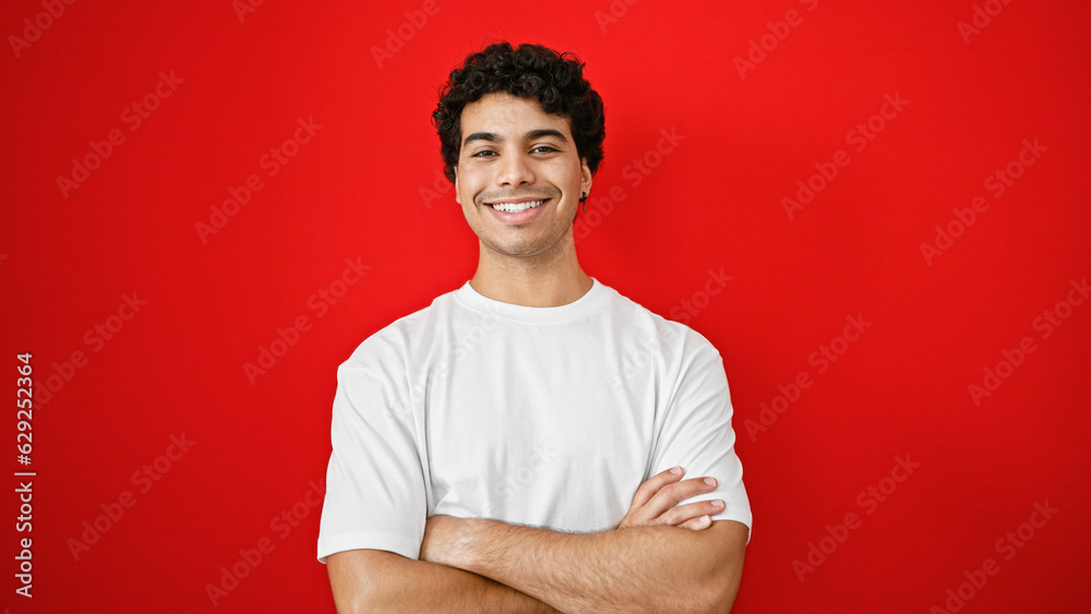 Young latin man smiling confident standing with arms crossed gesture over isolated red background Young latin man smiling confident standing with arms crossed gesture over isolated red background