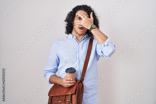 Hispanic man with curly hair drinking a cup of take away coffee peeking in shock covering face and eyes with hand, looking through fingers with embarrassed expression.