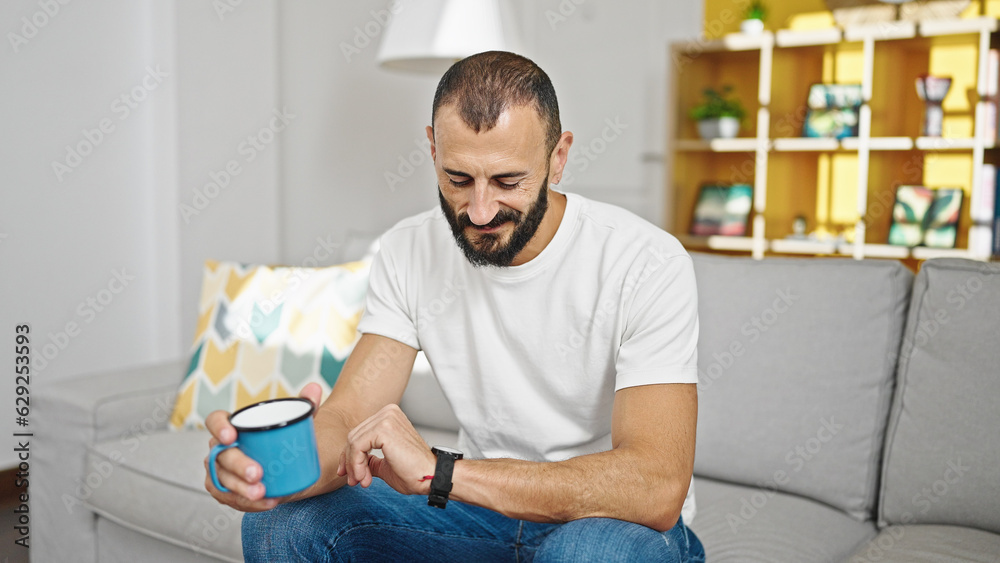 Young hispanic man drinking coffee looking watch at home