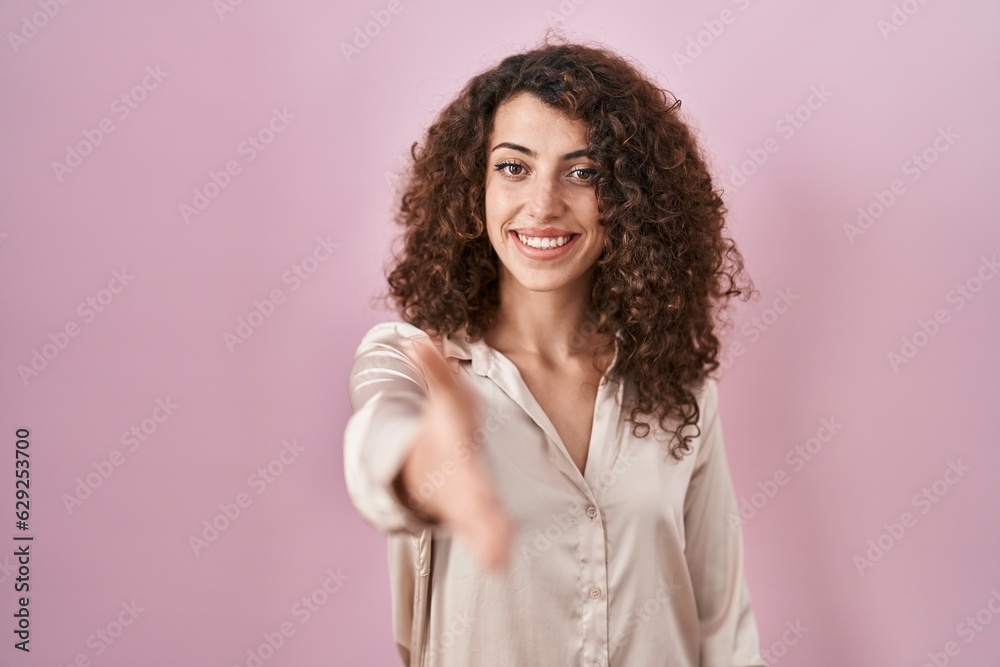 Hispanic woman with curly hair standing over pink background smiling friendly offering handshake as greeting and welcoming. successful business. Hispanic woman with curly hair standing over pink background smiling friendly offering handshake as greeting and welcoming. successful business.