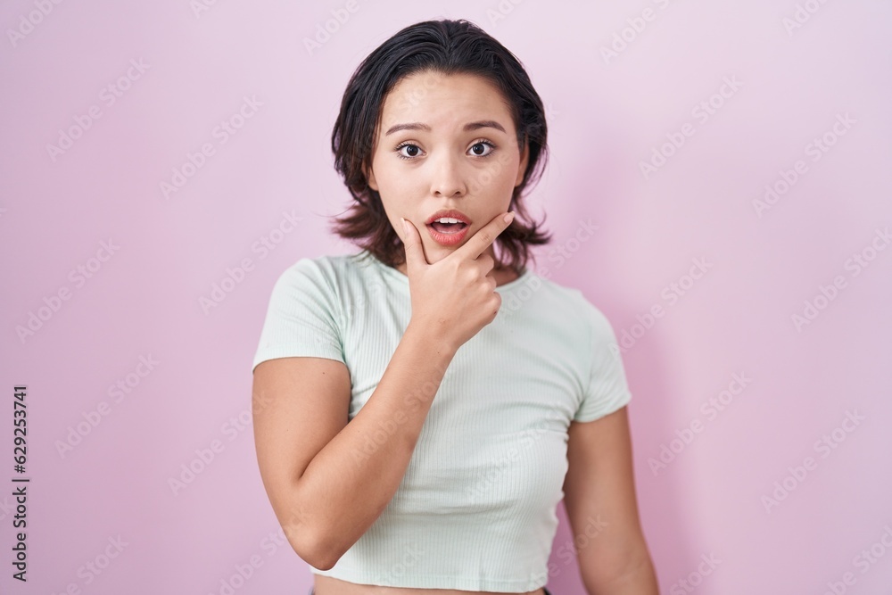 Hispanic young woman standing over pink background looking fascinated with disbelief, surprise and amazed expression with hands on chin Hispanic young woman standing over pink background looking fascinated with disbelief, surprise and amazed expression with hands on chin