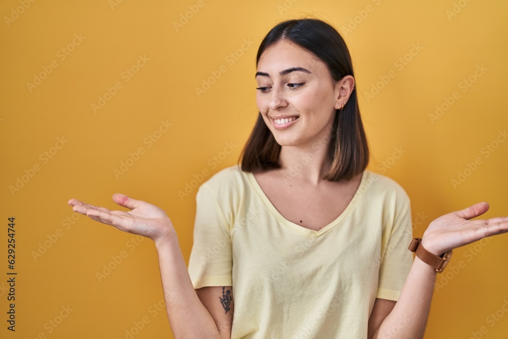 Hispanic girl wearing casual t shirt over yellow background smiling showing both hands open palms, presenting and advertising comparison and balance Hispanic girl wearing casual t shirt over yellow background smiling showing both hands open palms, presenting and advertising comparison and balance