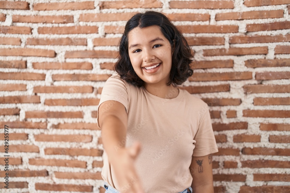 Young hispanic woman standing over bricks wall smiling friendly offering handshake as greeting and welcoming. successful business.