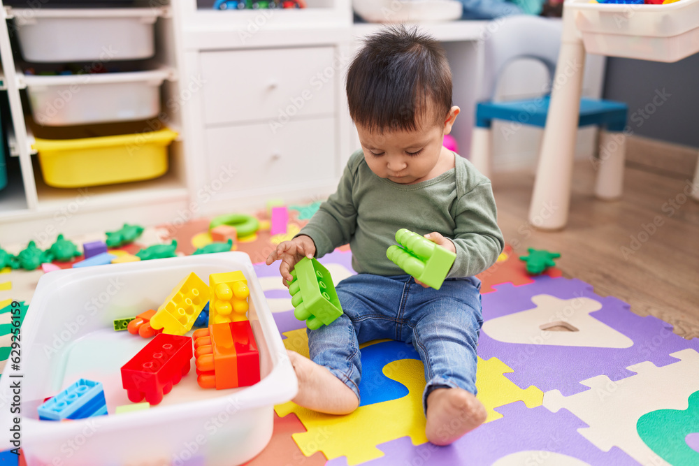 Fototapeta premium Adorable hispanic boy playing with construction blocks sitting on floor at kindergarten