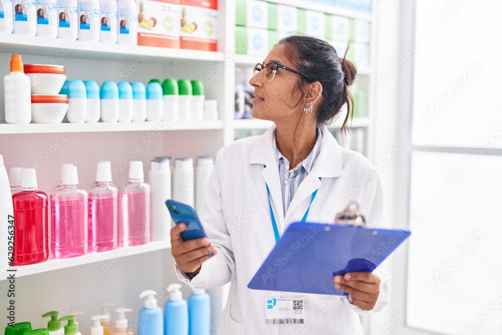 Young beautiful hispanic woman pharmacist using smartphone reading document at pharmacy