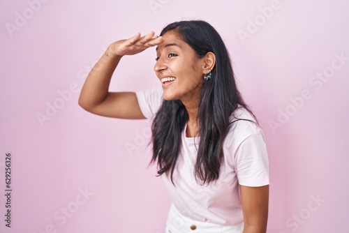 Young hispanic woman standing over pink background very happy and smiling looking far away with hand over head. searching concept.