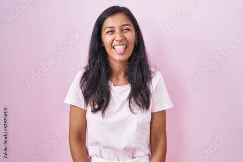 Young hispanic woman standing over pink background sticking tongue out happy with funny expression. emotion concept.