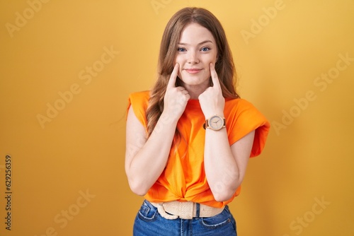 Caucasian woman standing over yellow background smiling with open mouth, fingers pointing and forcing cheerful smile