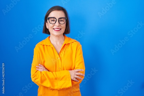 Middle age hispanic woman wearing glasses standing over blue background happy face smiling with crossed arms looking at the camera. positive person.