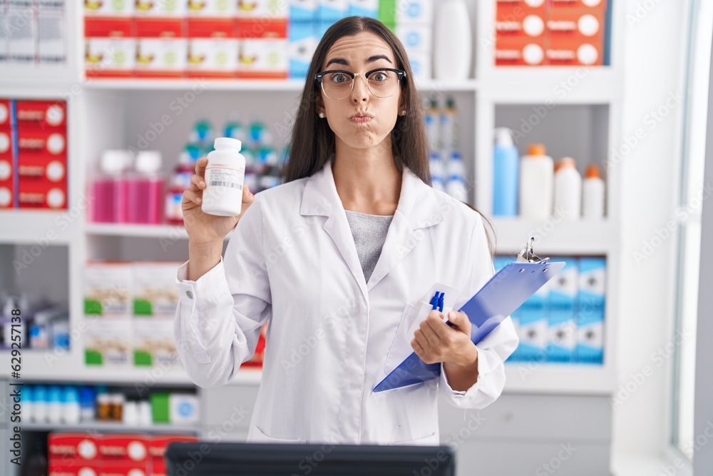 Young brunette woman working at pharmacy drugstore holding pills puffing cheeks with funny face. mouth inflated with air, catching air.
