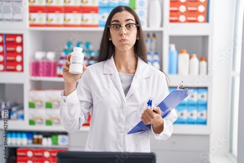 Young brunette woman working at pharmacy drugstore holding pills puffing cheeks with funny face. mouth inflated with air  catching air.