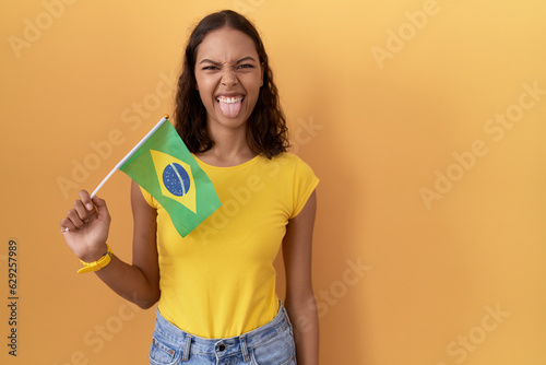 Young hispanic woman holding brazil flag sticking tongue out happy with funny expression. emotion concept.