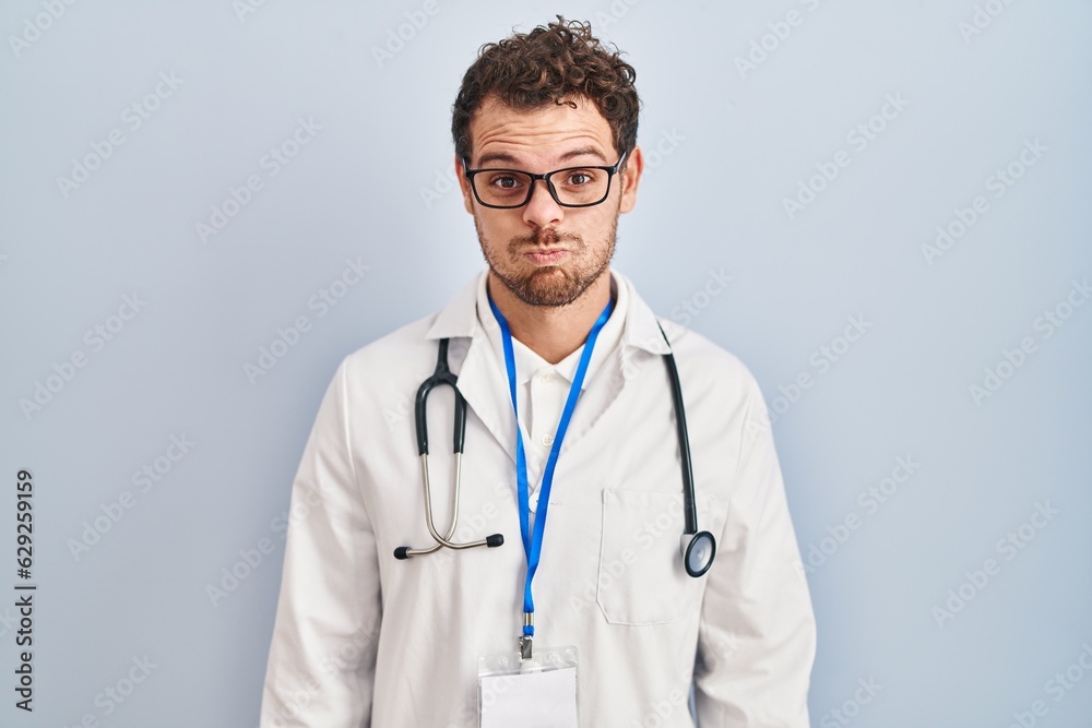 Young hispanic man wearing doctor uniform and stethoscope puffing cheeks with funny face. mouth inflated with air, crazy expression. Young hispanic man wearing doctor uniform and stethoscope puffing cheeks with funny face. mouth inflated with air, crazy expression.