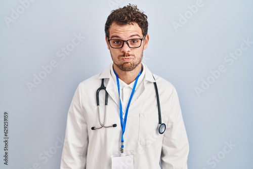 Young hispanic man wearing doctor uniform and stethoscope puffing cheeks with funny face. mouth inflated with air, crazy expression.