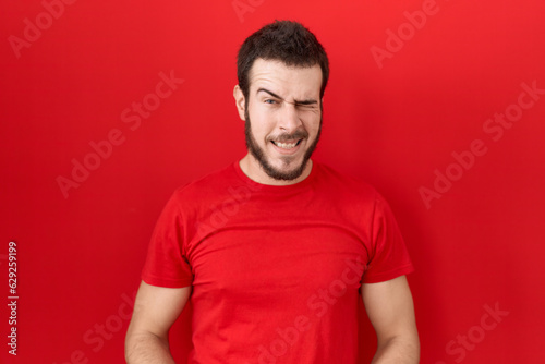 Young hispanic man wearing casual red t shirt winking looking at the camera with sexy expression, cheerful and happy face.