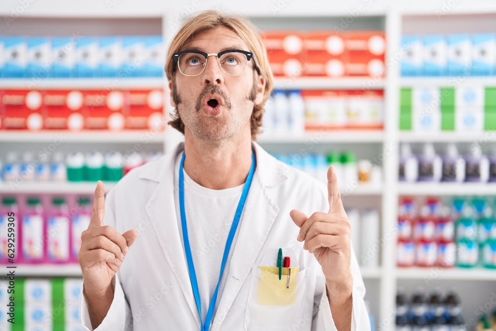 Caucasian man with mustache working at pharmacy drugstore amazed and surprised looking up and pointing with fingers and raised arms.