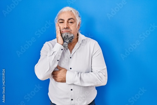 Middle age man with grey hair standing over blue background thinking looking tired and bored with depression problems with crossed arms.