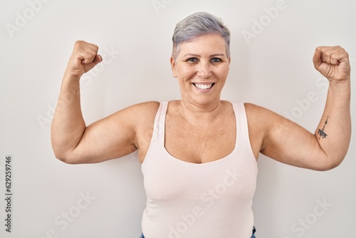 Middle age caucasian woman standing over white background showing arms muscles smiling proud. fitness concept.
